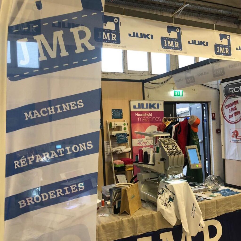 Visuel d'une flamme publicitaire pour JMR, affichant les services de machines, réparations et broderies, avec un aperçu d'un stand d'exposition montrant des machines à coudre et divers accessoires.