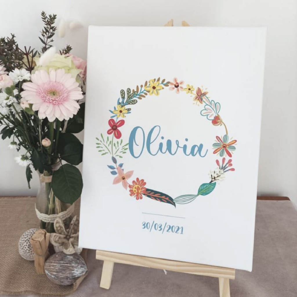 Visuel d'une impression sur toile posée sur un chevalet en bois, affichant le prénom 'Olivia' entouré d'une couronne de fleurs colorées et la date '30/03/2021', accompagnée d'un vase de fleurs à côté.