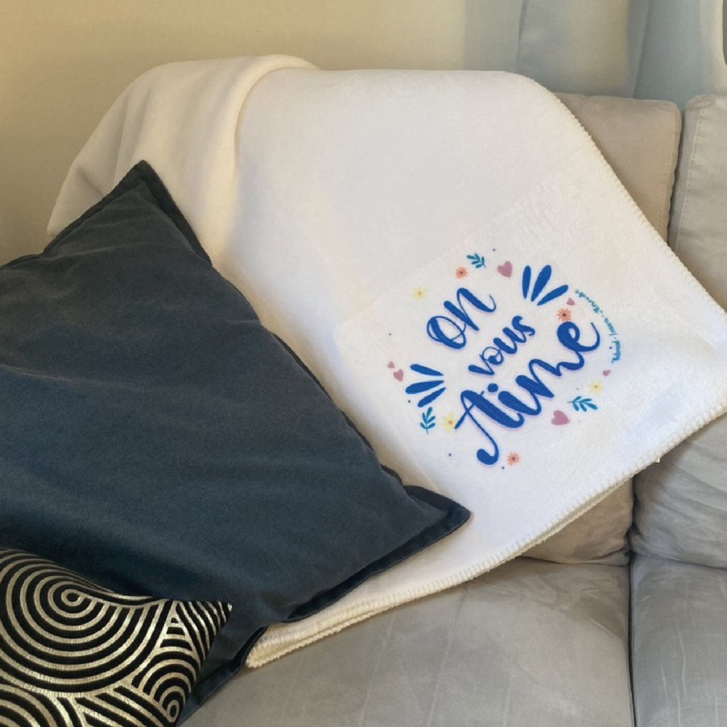 Visuel d'un plaid blanc posé sur un canapé avec des coussins, présentant une inscription colorée 'On vous aime' entourée de motifs floraux et de cœurs.
