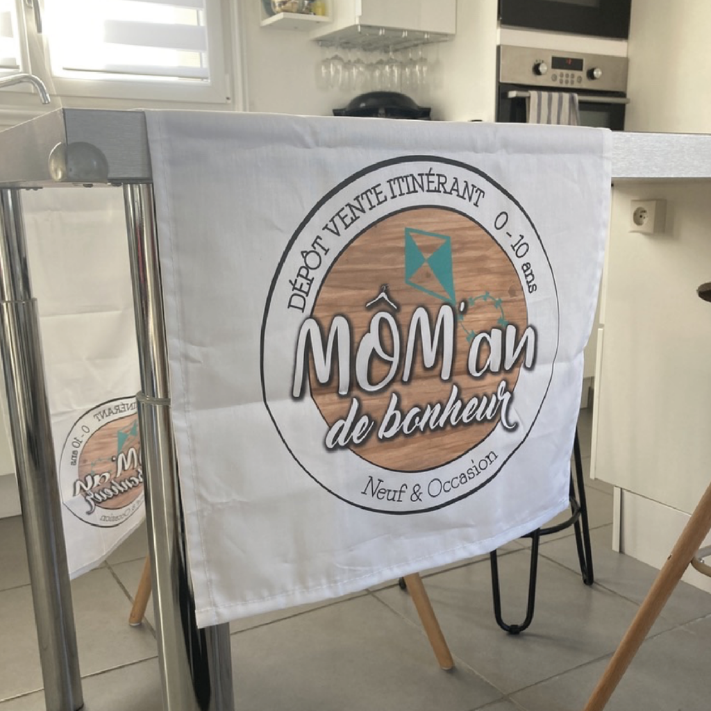 Visuel d'un chemin de table personnalisé pour un dépôt-vente itinérant nommé 'Môm'an de bonheur', spécialisé dans les articles pour nouveau-nés et occasions, avec un logo représentant un cerf-volant et des informations sur les horaires d'ouverture de 0 à 10 ans.