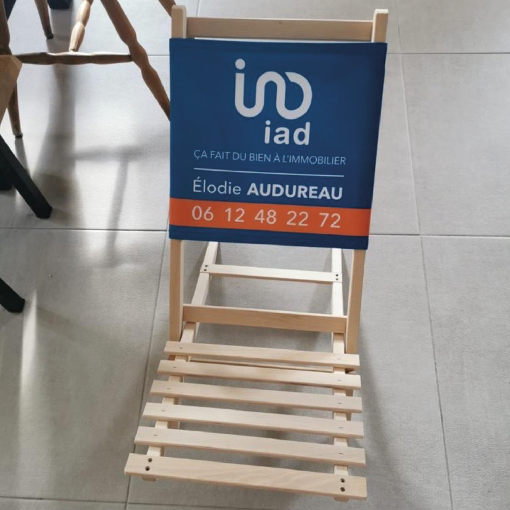 Visuel d'une chaise de plage en bois avec un panneau publicitaire pour iad, affichant le slogan 'Ça fait du bien à l'immobilier' et les informations de contact d'Élodie Audureau, agent immobilier, incluant un numéro de téléphone.