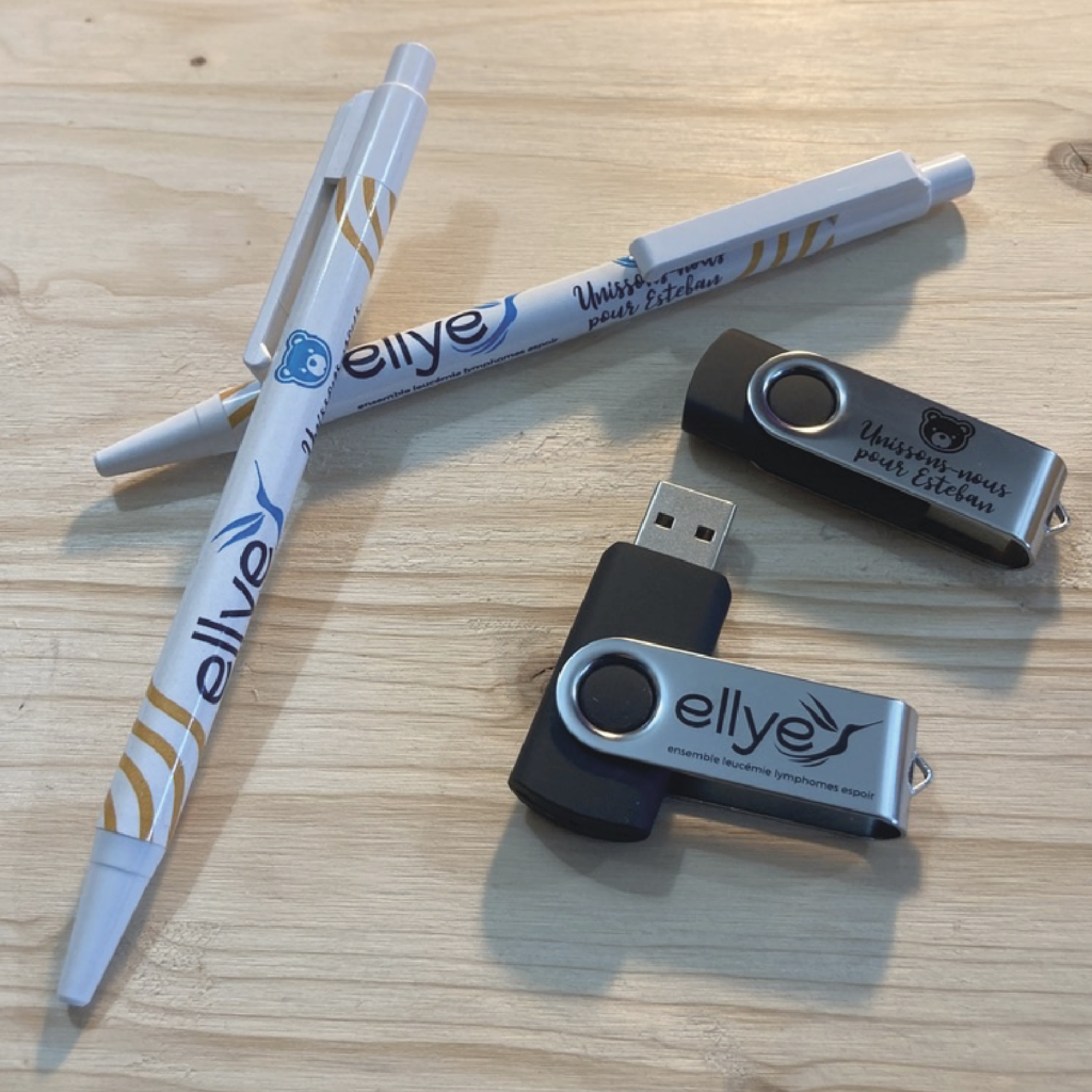 Visuel de stylos et de clés USB personnalisés pour Ellye, affichant le logo de l'entreprise et le slogan 'Unissons-nous pour l'Essentiel', posés sur une surface en bois.