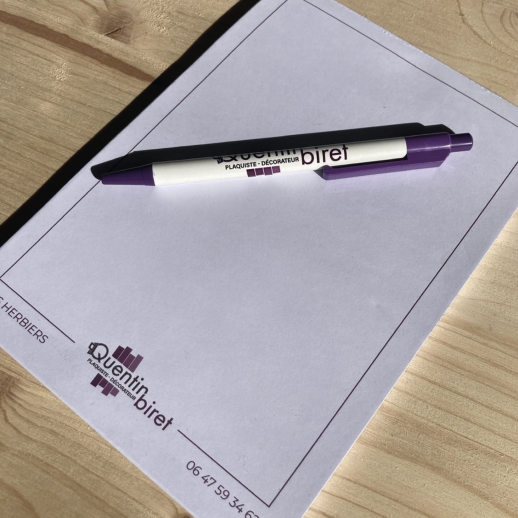 Visuel d'un stylo et d'un bloc-notes personnalisés pour Quentin Biret, plaquiste et décorateur, affichant le logo de l'entreprise et un numéro de téléphone, posés sur une surface en bois.