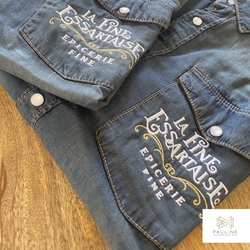 Deux chemises en tissu denim avec des broderies élégantes sur les poches de poitrine, affichant le texte 'La Fine Epicerie Fine' en lettres cursives blanches et dorées, posées sur une surface en bois.