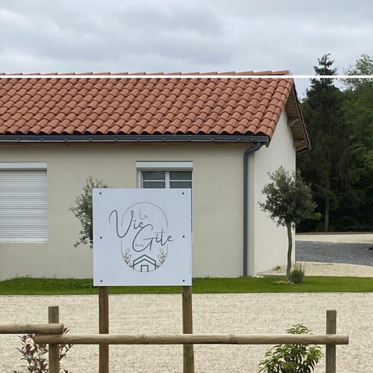 Panneau en dibond installé devant une maison avec un toit en tuiles rouges, affichant le texte 'La Vie en Gîte' avec un logo de maison, entouré d'un petit jardin bien entretenu et d'une clôture en bois.