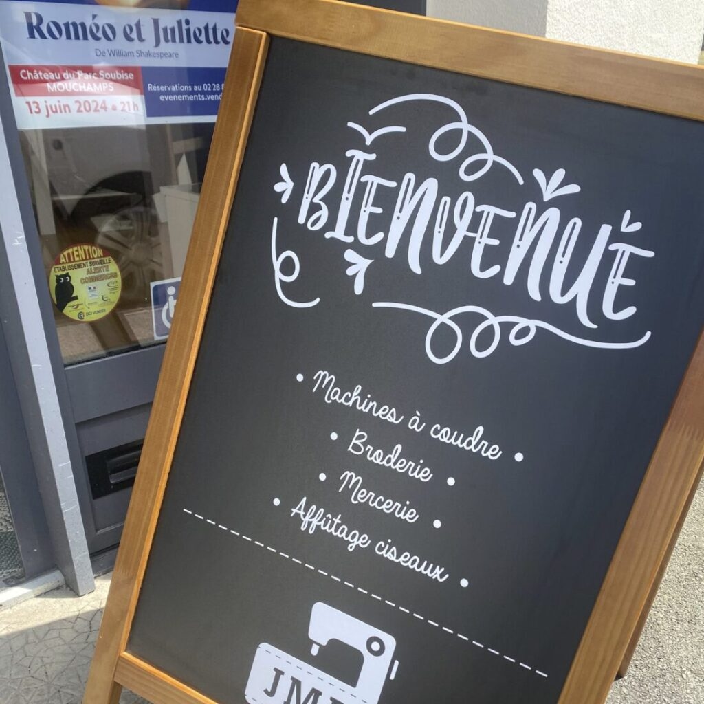 Tableau en ardoise avec cadre en bois affichant 'Bienvenue' en écriture stylisée, listant les services offerts : machines à coudre, broderie, mercerie, et affûtage de ciseaux, placé devant le magasin JMR.