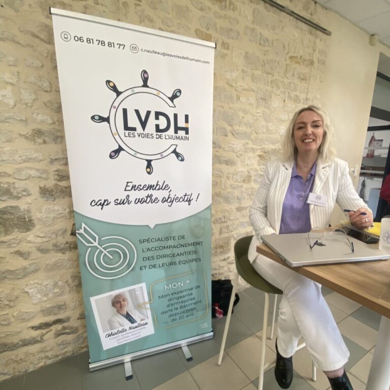 Une femme souriante, assise à une table avec un ordinateur portable, à côté d'un roll-up publicitaire pour LVDH - Les Voies de l'Humain, spécialiste de l'accompagnement des dirigeants et de leurs équipes, avec plus de 20 ans d'expérience en entreprise.